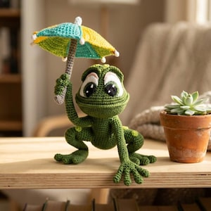 May include: A green crocheted frog figurine with large white and black eyes, holding a small, multi-colored crocheted umbrella with teal, yellow, and white segments. The frog has a textured surface and is positioned next to a small potted succulent.