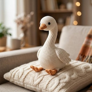 May include: A cream-colored crocheted goose plush toy with a long neck, small black eyes, and an orange beak and feet. The goose is crafted with a visible yarn stitch pattern and is positioned sitting upright on a textured cream-colored knit pillow.