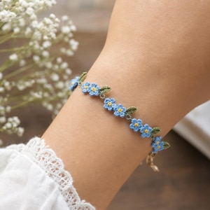 Op de afbeelding: Een delicate armband met een reeks kleine, lichtblauwe gehaakte vergeet-mij-nietjes met felgele hartjes, afgewisseld met kleine groene gehaakte blaadjes, verbonden door een dunne metalen ketting met een sluiting.