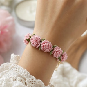 May include: A bracelet featuring a series of small, light pink crocheted rose blossoms connected by tiny green crocheted leaves. The floral design is crafted from fine thread, creating a delicate texture. A thin gold-toned metal chain is visible.