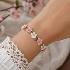 以下が含まれることがあります： 淡いピンクと白の小さなかぎ針編みの花モチーフが連なり、小さな緑の葉がアクセントになった繊細なブレスレット。花のチェーンは、小さな留め具が付いた細いゴールドトーンの金属チェーンにつながっています。