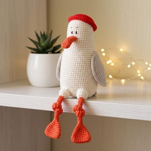 May include: A handmade crochet seagull toy featuring a cream-colored body, a bright red hat, and a matching orange beak and webbed feet. The bird has gray wings and is designed to sit upright with its long, dangling legs hanging over the edge.