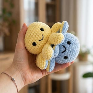 Op de afbeelding: Een klein, handgemaakt gehaakt knuffeltje met twee met elkaar verbonden inktvisfiguren. De ene inktvis is felgeel en de andere is lichtblauw. Beide hebben ronde koppen, kleine zwarte plastic ogen en geborduurde lachende mondjes.