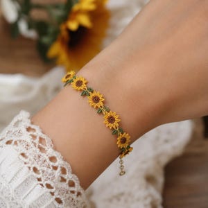 May include: A delicate bracelet featuring a series of small, intricately stitched sunflowers with bright yellow petals, dark brown centres, and tiny green leaves. The floral chain is connected to a thin metal link extension chain with a small clasp.