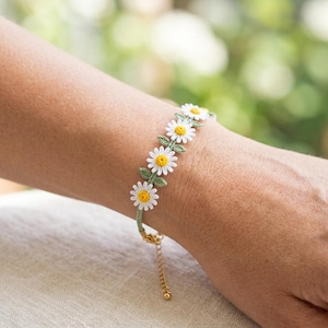 May include: A bracelet featuring a series of white daisy flowers with yellow centers and green leaves, crafted from thread. The floral design is connected by a thin green cord and includes a gold-toned metal chain extension with a small round clasp.