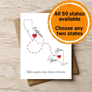 May include: A white greeting card featuring black outlines of California and New York, each marked with a red heart. A dashed red line connects the two states. The text reads: "California", "New York", and "Miles apart, but close at heart."