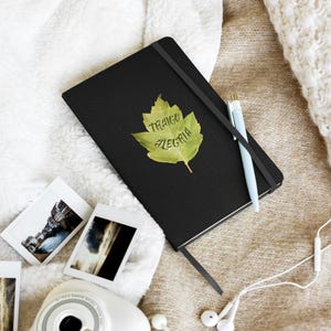 Peut inclure: Un carnet noir avec une bande élastique de fermeture et un stylo bleu clair posé dessus. Un motif de feuille verte est imprimé sur la couverture avec le texte "TRAIGO ALEGRIA" écrit à l'encre noire au centre de la surface de la feuille.