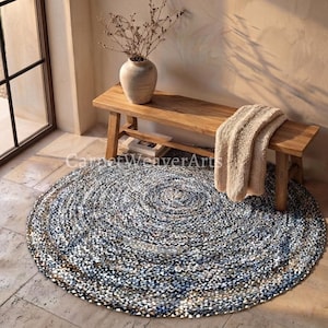 May include: A round, braided area rug featuring a multi-tonal pattern of blue, grey, white, and tan fibers. The rug is positioned on a stone floor beneath a wooden bench that holds a beige knit throw blanket and a ceramic vase with dried branches.