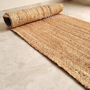 Puede incluir: Una alfombra rectangular hecha de fibras de yute de color tostado natural, parcialmente desenrollada para revelar una textura trenzada gruesa. La superficie presenta un patrón tejido consistente en un tono marrón claro, con el texto CarpetWeaverArts.