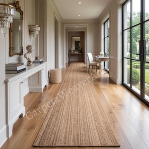 May include: A long, rectangular runner rug made of natural tan woven fibres, positioned centrally on a light-toned hardwood floor in a hallway. The rug features a consistent, textured weave pattern that runs the entire length of the floor space.