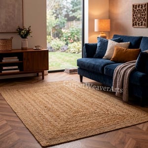 Puede incluir: Una alfombra rectangular hecha de fibras de yute natural de color tostado con una textura trenzada y un diseño de borde distintivo. La alfombra está colocada sobre un suelo de madera en una habitación con un sofá azul oscuro y una consola.