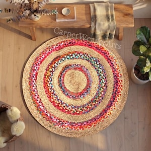 Puede incluir: Una alfombra redonda de yute trenzado que presenta círculos concéntricos de fibras de color tostado natural intercalados con tiras de tela vibrantes y multicolores en tonos rosa, azul, amarillo y morado, creando un patrón circular texturizado.