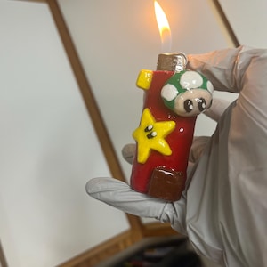 Pode incluir: Um isqueiro vermelho com uma estrela amarela tridimensional e um cogumelo verde e branco com pintas fixado na frente. O isqueiro está aceso, produzindo uma pequena chama laranja no topo. A superfície do isqueiro tem um acabamento brilhante.