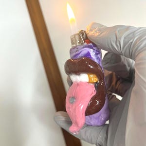 May include: A purple lighter featuring a sculpted design of brown lips, white teeth with a gold tooth, and a long pink tongue with a small silver stud. The lighter is lit, producing a visible yellow flame at the top metal ignition mechanism.