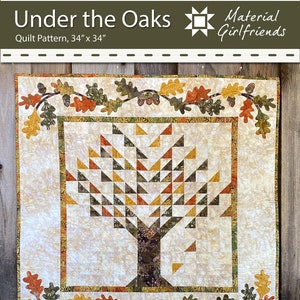 以下が含まれることがあります： マテリアル・ガールフレンドによる「Under the Oaks」というタイトルのキルトパターン。キルトは34インチ×34インチで、茶色の幹と緑、黄、オレンジの葉を持つ木が描かれています。木は緑、黄、オレンジの色のオークの葉の縁取りで囲まれています。