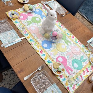 Puede incluir: Una figurita de conejo de cer&aacute;mica blanca se encuentra sobre una mesa con un mantel de mesa colorido con tem&aacute;tica de Pascua. El mantel de mesa presenta un patr&oacute;n de conejos y huevos. Tambi&eacute;n hay vasos, platos y otras decoraciones en la mesa.