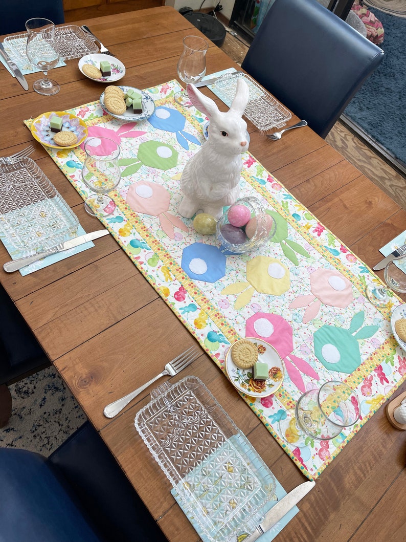 Puede incluir: Una figurita de conejo de cer&aacute;mica blanca se encuentra sobre una mesa con un mantel de mesa colorido con tem&aacute;tica de Pascua. El mantel de mesa presenta un patr&oacute;n de conejos y huevos. Tambi&eacute;n hay vasos, platos y otras decoraciones en la mesa.