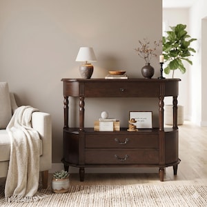 May include: A dark brown wooden console table featuring a curved top, one upper drawer with a small knob, and two lower drawers with ornate metal pulls. The table is supported by four turned legs and includes a lower shelf for additional storage space.
