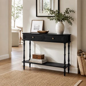 May include: A black console table featuring two drawers with small gold knobs, slender turned legs, and a lower shelf. The table surface holds a white ceramic vase with green foliage and a wooden bowl, while the lower shelf holds books.