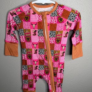 Op de afbeelding: Een baby-rompertje met lange mouwen en een patchworkpatroon van roze, bruine en zwart-witte dierenprints. Het ontwerp bevat koeienschedels, cowboylaarzen en biohazard-symbolen. Het heeft een bruine kraag, mouwboorden en een ritssluiting.