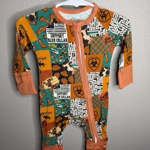 Op de afbeelding: Een baby-slaappak met lange mouwen en ritssluiting, voorzien van een patchwork-print van cowboylaarzen, gereedschap en de tekst SUPPORT BLUE COLLAR en THE WORLD NEEDS MORE COWBOYS in de kleuren oranje, groen en wit.