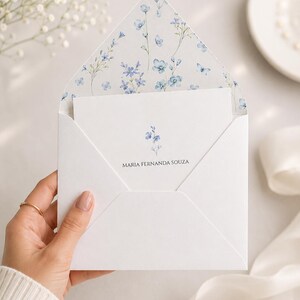 Pode incluir: Um envelope branco com aba pontiaguda apresentando um padrão floral azul no forro interno. Um cartão branco está inserido dentro, impresso com uma pequena ilustração de flor azul e o nome MARIA FERNANDA SOUZA em letras maiúsculas pretas.