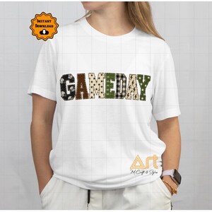 Op de afbeelding: Een wit T-shirt met korte mouwen en het woord GAMEDAY in grote blokletters op de borst. Elke letter heeft een uniek patroon, waaronder footballhelmen, bruin ruitpatroon, sterren, een groen speelveldgrid en bliksemschichten.