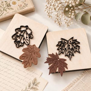 May include: Two square light wood blocks each featuring a black metal leaf-shaped cookie cutter and a corresponding brown textured leaf-shaped cutout. The cutters display intricate vein patterns and small circular details within the leaf outlines.