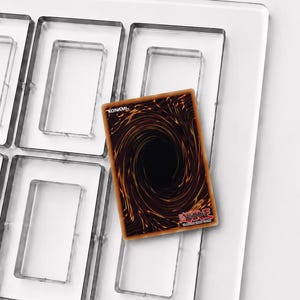 Puede incluir: Un marco de exhibición de acrílico transparente con múltiples ranuras rectangulares para tarjetas coleccionables. Una carta de Yu-Gi-Oh! se coloca sobre una ranura, mostrando el diseño posterior con un patrón de vórtice oscuro y el logo.