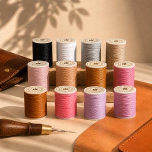 Peut inclure: Une collection de douze bobines de fil cylindriques de différentes couleurs, dont noir, blanc, gris, beige, rose, marron et lavande. Chaque bobine présente une étiquette circulaire sur le dessus avec une icône d'étoile et du texte. Un poinçon en bois est visible.