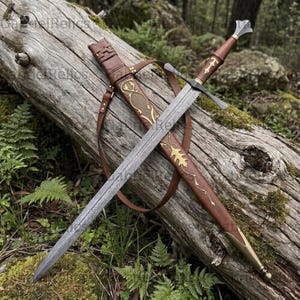 Può includere: Una spada a doppio taglio con lama in acciaio damasco a motivi, una guardia trasversale e un fodero in pelle marrone con accenti in metallo dorato, tra cui un dettaglio a forma di foglia e una cinghia in pelle per il trasporto.