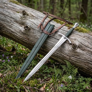 Puede incluir: Una espada de acero de doble filo con empuñadura verde oscuro y una vaina a juego. La vaina está sujeta a un arnés de cuero marrón con hebillas y correas de metal. La espada presenta una guarda plateada y una hoja puntiaguda.
