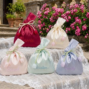 Pode incluir: Cinco bolsas de cetim com fecho de cordão nas cores bordô, creme, rosa claro, verde menta e azul claro. Cada bolsa apresenta um nome bordado em letra cursiva e um fecho de cordão com extremidades em formato de tulipa.
