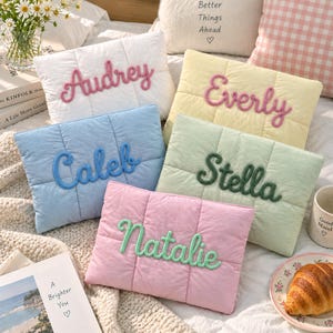 May include: Five rectangular quilted pillows in white, yellow, blue, light green, and pink. Each pillow features a name embroidered in a raised, cursive yarn script: Audrey, Everly, Caleb, Stella, and Natalie. The pillows have a soft, padded texture.