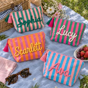 May include: Four rectangular zippered pouches with vertical stripes in various color combinations. Each pouch features a personalized name in raised, textured script lettering and a matching tassel attached to the gold-tone zipper pull.