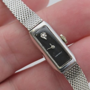 May include: A silver-tone rectangular wristwatch with a black dial, a single diamond accent at the twelve o'clock position, silver hands, and a textured mesh metal strap. The dial features the text Longines in white lettering.