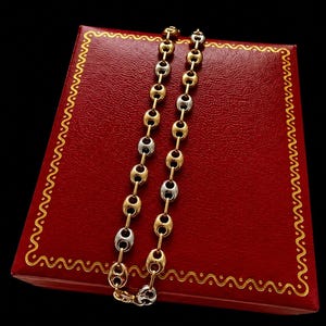 May include: A two-tone chain necklace featuring alternating oval-shaped links in polished yellow gold and silver-toned metal. The chain is displayed on a rectangular red box with a textured surface and a decorative gold-colored border pattern.