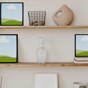 May include: A display of decorative items on wooden shelves. The shelves hold framed landscape art, a candle, a wire basket, a vase, a mug, a decanter, and books. The art features a cloud and green hills.