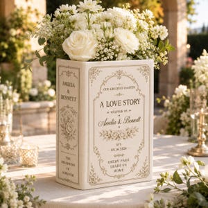 May include: A cream-colored ceramic book-shaped vase featuring gold-toned text that reads A LOVE STORY, WRITTEN BY AMELIA & BENNETT, EST. 08.16.2026, EVERY PAGE LEADS US HOME, and AMELIA & BENNETT on the spine, filled with white roses and baby's breath.