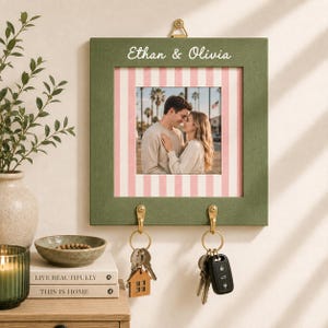 Könnte beinhalten: Ein quadratischer, olivgrüner Schlüsselhalter aus Holz zur Wandmontage mit einem zentralen Foto eines Paares. Der Rahmen hat weiße und rosa vertikale Streifen, die Namen Ethan & Olivia oben und zwei Messinghaken für Schlüssel.