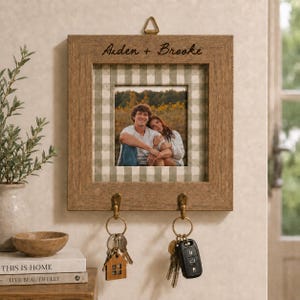 May include: A square wooden wall-mounted key holder featuring a light brown frame with the engraved text Aiden + Brooke. The center displays a photograph of two people against a sage green and white gingham background, with two metal hooks for keys.