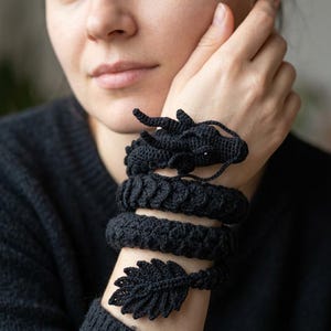 May include: A black crocheted dragon bracelet that wraps around the wrist. The design features a detailed dragon head with horns and whiskers at one end, a textured scaly body, and a pointed, leaf-shaped tail at the other end of the accessory.