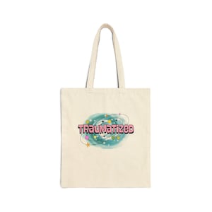 May include: A beige canvas tote bag featuring a central graphic of a teal swirling galaxy with small colorful stars and planets. The word TRAUMATIZED is printed in large, bold, pink block letters with a black outline across the center of the design.