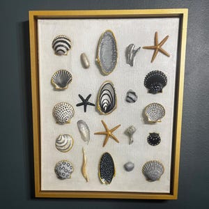 May include: A rectangular wall art piece featuring a collection of various seashells and starfish arranged on a white textured background, all enclosed within a gold frame. The shells display black, white, and metallic gold painted patterns.