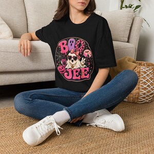 May include: A black t-shirt featuring a circular graphic with the text BOO JEE in pink. The design includes a ghost wearing heart-shaped sunglasses and a pink bow, holding a drink, surrounded by a spider web, a pumpkin, and a haunted castle.