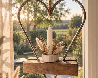 May include: A hanging heart-shaped metal frame suspended by a jute rope. A rectangular wooden block sits at the base of the heart, supporting a white fluted candle holder with a white pillar candle surrounded by dried beige grasses and foliage.