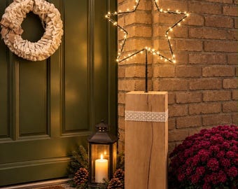 May include: A light-up star decoration mounted on a tall, rectangular wooden block base with a white lace ribbon, positioned next to a black metal lantern with a lit candle and a wreath on a dark green door.