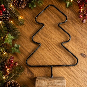 May include: A black metal wire frame in the shape of a Christmas tree, featuring a textured, twisted surface. The wire tree is mounted on a rectangular, light-colored wooden base, providing a stable foundation for the decorative structure.