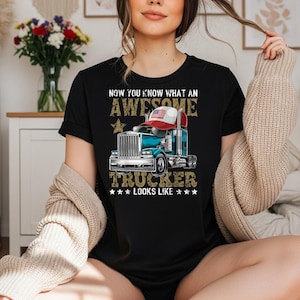 Puede incluir: Una camiseta negra de manga corta con el gráfico de un camión de color azul verdoso que lleva una gorra de béisbol roja, blanca y azul. El texto dice: NOW YOU KNOW WHAT AN AWESOME TRUCKER LOOKS LIKE. El diseño incluye estrellas doradas y letras desgastadas.