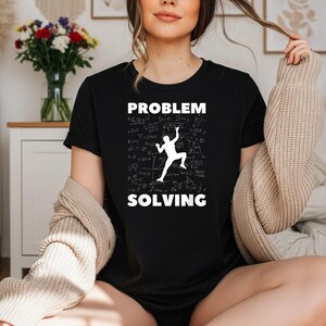以下が含まれることがあります： 白いグラフィックプリントが施された黒の半袖Tシャツ。デザインには太字の大文字で「PROBLEM SOLVING」というテキストがあり、背景にはクライミングをする人のシルエットと様々な数学の方程式が描かれています。